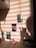 A wall with several pictures attached, illuminated by sunlight filtering through blinds, casting striped shadows. A silhouette of a person is visible, making a peace sign. Below, a few sticky notes and a stack of colored paper are placed on a surface.