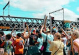 A vibrant crowd participates in an outdoor event, celebrating with colorful attire and rainbow-themed decorations. Many individuals wear rainbow hats and carry long, rainbow-colored inflatables, amidst a backdrop of an urban environment with a bridge and train tracks. The atmosphere appears festive and lively.