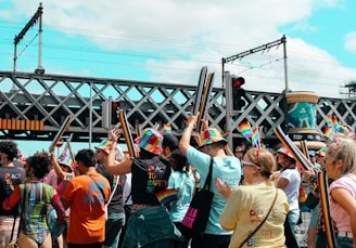 A vibrant crowd participates in an outdoor event, celebrating with colorful attire and rainbow-themed decorations. Many individuals wear rainbow hats and carry long, rainbow-colored inflatables, amidst a backdrop of an urban environment with a bridge and train tracks. The atmosphere appears festive and lively.