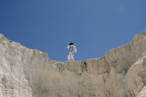 An astronaut stands at the edge of a rocky cliff against a clear blue sky, appearing to look into the distance.