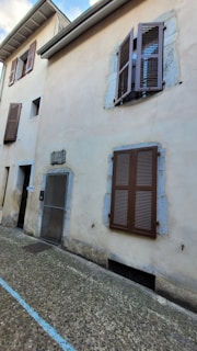 A beige-colored building with brown wooden shutters and a stone frame around the windows. The building has an entrance with a metal mesh door and a small plaque next to it. The ground is paved with stones and a blue line runs along it.