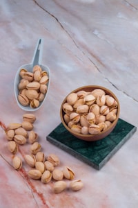 A collection of pistachios is displayed, with some contained in a small white scoop and others in a wooden bowl. Several pistachios are scattered across a marble surface, which has a blend of light pink and gray tones. The bowl is placed on a green marble square, contrasting the background.