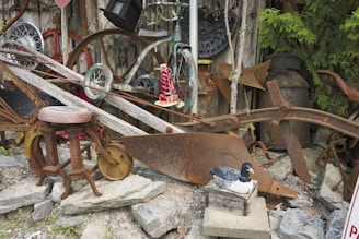 A collection of rustic, weathered objects including old wheels, metal implements, and tools are arranged against a wooden wall. There is a wooden stool with a metal seat, an old plow, and various other vintage farm equipment. A decorative wooden bird is placed on a box in the foreground.