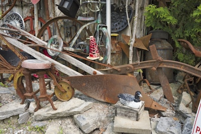 A collection of rustic, weathered objects including old wheels, metal implements, and tools are arranged against a wooden wall. There is a wooden stool with a metal seat, an old plow, and various other vintage farm equipment. A decorative wooden bird is placed on a box in the foreground.