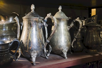 An arrangement of vintage silver teapots and pitchers is set on a wooden shelf, showing signs of tarnish and age with intricate detailing on handles and spouts illuminated by soft sunlight.