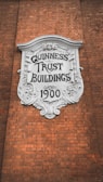 A decorative plaque with ornate detailing is mounted on a brick wall. The white plaque has 'GUINNESS TRUST BUILDINGS 1900' inscribed on it in bold, stylized letters.