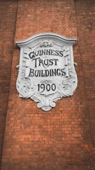 A decorative plaque with ornate detailing is mounted on a brick wall. The white plaque has 'GUINNESS TRUST BUILDINGS 1900' inscribed on it in bold, stylized letters.