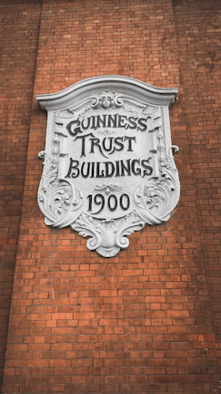 A decorative plaque with ornate detailing is mounted on a brick wall. The white plaque has 'GUINNESS TRUST BUILDINGS 1900' inscribed on it in bold, stylized letters.