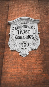 A decorative plaque with ornate detailing is mounted on a brick wall. The white plaque has 'GUINNESS TRUST BUILDINGS 1900' inscribed on it in bold, stylized letters.