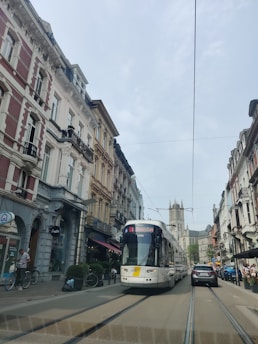 A tram is moving down a street lined with historic European architecture. The buildings feature classic facades with balconies and shops at ground level. People can be seen near the tram and bicycles are parked along the sidewalk. A church with a tall spire is visible in the background.