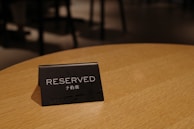 A small black sign that reads 'RESERVED' in English and additional text in another language is placed upon a wooden table. The background is softly blurred, emphasizing the sign as the focal point.