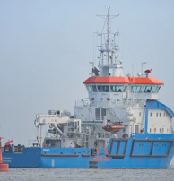 A large blue and white ship with orange accents is positioned on calm waters. The ship has a complex array of antennas and equipment on its upper deck. A small red buoy is floating nearby with a flag attached.