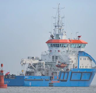 A large blue and white ship with orange accents is positioned on calm waters. The ship has a complex array of antennas and equipment on its upper deck. A small red buoy is floating nearby with a flag attached.