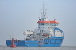 A large blue and white ship with orange accents is positioned on calm waters. The ship has a complex array of antennas and equipment on its upper deck. A small red buoy is floating nearby with a flag attached.