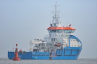 A large blue and white ship with orange accents is positioned on calm waters. The ship has a complex array of antennas and equipment on its upper deck. A small red buoy is floating nearby with a flag attached.