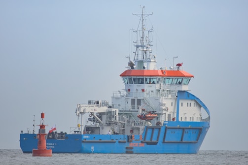 A large blue and white ship with orange accents is positioned on calm waters. The ship has a complex array of antennas and equipment on its upper deck. A small red buoy is floating nearby with a flag attached.