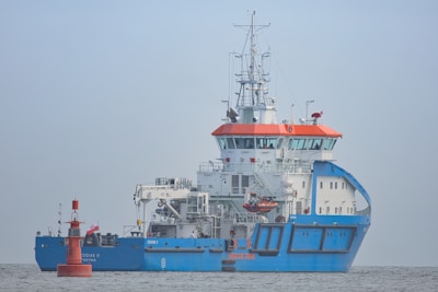 A large blue and white ship with orange accents is positioned on calm waters. The ship has a complex array of antennas and equipment on its upper deck. A small red buoy is floating nearby with a flag attached.