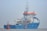 A large blue and white ship with orange accents is positioned on calm waters. The ship has a complex array of antennas and equipment on its upper deck. A small red buoy is floating nearby with a flag attached.