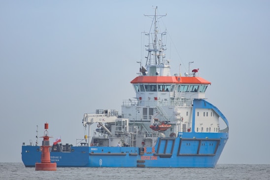 A large blue and white ship with orange accents is positioned on calm waters. The ship has a complex array of antennas and equipment on its upper deck. A small red buoy is floating nearby with a flag attached.