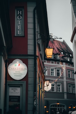 A narrow street featuring a building with a bright sign for Casa Italia, a hotel and pizzeria. The area is dimly lit, with ambient lighting from street lamps, creating a cozy atmosphere. Adjacent buildings show classic architecture with decorative facades.