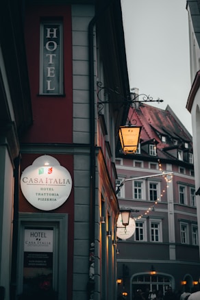 A narrow street featuring a building with a bright sign for Casa Italia, a hotel and pizzeria. The area is dimly lit, with ambient lighting from street lamps, creating a cozy atmosphere. Adjacent buildings show classic architecture with decorative facades.
