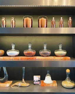 Shelves display a collection of copper drink shakers and ice buckets with glass containers filled with various colored sands or powders. The containers are arranged on square stone bases, with some creatively shaped for aesthetic appeal.