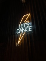 Neon sign displaying the words 'Let’s Dance' with a stylized lightning bolt design. The sign emits a bright glow against a dark, textured backdrop.