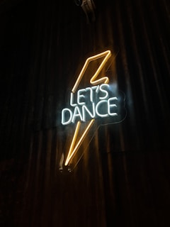 Neon sign displaying the words 'Let’s Dance' with a stylized lightning bolt design. The sign emits a bright glow against a dark, textured backdrop.