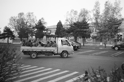 A group of people sitting in the back of a light truck moving along a road near a zebra crossing. The background includes trees and a building with a fence. Several cars are parked or driving nearby.