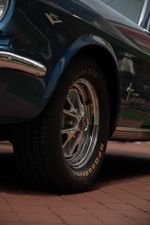 A close-up of a vintage car's front tire and wheel, featuring chrome details and radial tires with 'BFGoodrich' branding. The car is parked on a brick surface, and part of the blue metallic body with chrome trimming is visible.