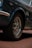 A close-up of a vintage car's front tire and wheel, featuring chrome details and radial tires with 'BFGoodrich' branding. The car is parked on a brick surface, and part of the blue metallic body with chrome trimming is visible.