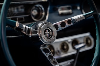 A close-up view of a classic Ford Mustang steering wheel, featuring the iconic emblem at the center surrounded by chrome details. The dashboard with circular vents and a gear shift is also visible in the background, contributing to the vintage car interior aesthetic.