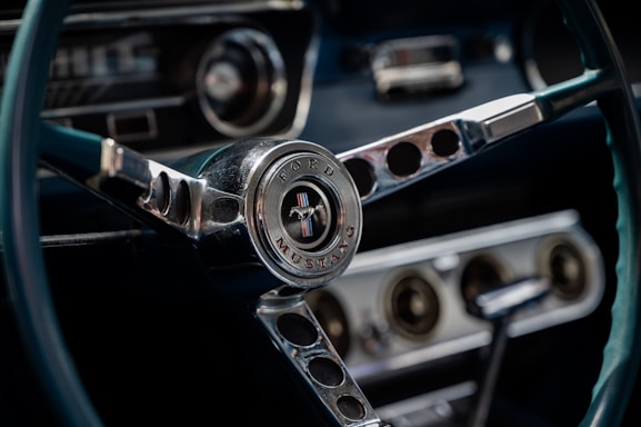A close-up view of a classic Ford Mustang steering wheel, featuring the iconic emblem at the center surrounded by chrome details. The dashboard with circular vents and a gear shift is also visible in the background, contributing to the vintage car interior aesthetic.