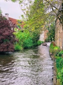 A serene waterway flows through a lush, green environment, bordered by vibrant trees and brick buildings. A small boat with several people on board navigates the waterway, creating a peaceful and communal atmosphere.