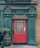 A pub entrance with a vibrant red door surrounded by green paneling and brickwork. Above the door, there is signage reading 'The Flavour Of Ireland & The British Isles.' Two decorative red stag head sculptures are mounted on either side of the sign. In front of the door, a wooden barrel with a flower pot and a black bollard can be seen.