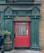 A pub entrance with a vibrant red door surrounded by green paneling and brickwork. Above the door, there is signage reading 'The Flavour Of Ireland & The British Isles.' Two decorative red stag head sculptures are mounted on either side of the sign. In front of the door, a wooden barrel with a flower pot and a black bollard can be seen.