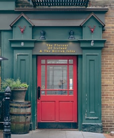 A pub entrance with a vibrant red door surrounded by green paneling and brickwork. Above the door, there is signage reading 'The Flavour Of Ireland & The British Isles.' Two decorative red stag head sculptures are mounted on either side of the sign. In front of the door, a wooden barrel with a flower pot and a black bollard can be seen.