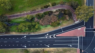 An aerial view of a multi-lane road intersecting with a bike path surrounded by greenery. The road features clear lane markings and directional arrows. The bike path meanders through a grassy area with several trees and a small stream visible.