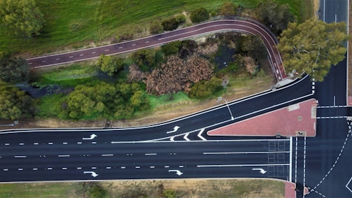 An aerial view of a multi-lane road intersecting with a bike path surrounded by greenery. The road features clear lane markings and directional arrows. The bike path meanders through a grassy area with several trees and a small stream visible.