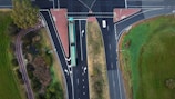 An aerial view of a T-shaped intersection with multiple traffic lanes. A bus is traveling along the main road, and a white car is seen approaching the intersection. Surrounding the roads are areas of green grass and trees, and there are designated pedestrian pathways.