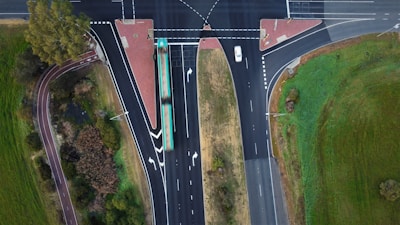 An aerial view of a T-shaped intersection with multiple traffic lanes. A bus is traveling along the main road, and a white car is seen approaching the intersection. Surrounding the roads are areas of green grass and trees, and there are designated pedestrian pathways.