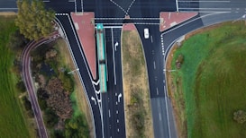An aerial view of a T-shaped intersection with multiple traffic lanes. A bus is traveling along the main road, and a white car is seen approaching the intersection. Surrounding the roads are areas of green grass and trees, and there are designated pedestrian pathways.