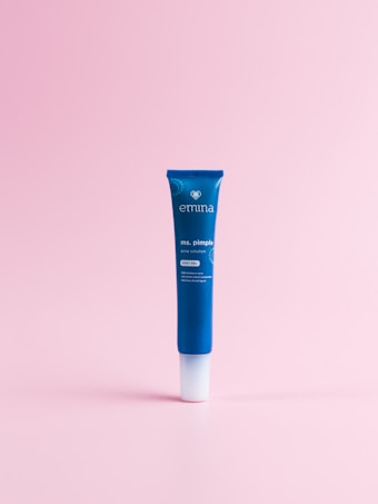 A blue tube labeled 'emina ms. pimple spot gel' is standing upright on a soft pink background. The design includes a small heart symbol and text detailing its purpose as an acne solution.