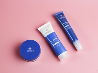 A flat lay of three skincare products on a soft pink background. The items include a small round container and two cylindrical tubes, all featuring a blue color with white accents. The brand name 'emina' is visible on the packaging.