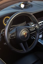 A detailed view of a luxury car's steering wheel, featuring a prominent logo at the center. The dashboard includes a circular instrument cluster with a yellow accent, and various control buttons on the steering wheel. The interior showcases high-quality materials with visible stitching.