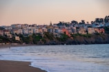 A coastal scene featuring a row of colorful houses perched on a cliff overlooking the ocean. The shoreline curves gently, with waves lapping at the sandy beach below. A few people can be seen walking along the water's edge, and there is a peaceful ambiance created by the clear sky and calm sea.