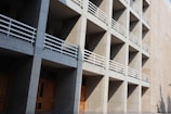 A modern architectural structure characterized by a grid of concrete modules, each with wooden doors and white railings, casting sharp shadows from sunlight.