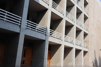 A modern architectural structure characterized by a grid of concrete modules, each with wooden doors and white railings, casting sharp shadows from sunlight.