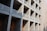 A modern architectural structure characterized by a grid of concrete modules, each with wooden doors and white railings, casting sharp shadows from sunlight.