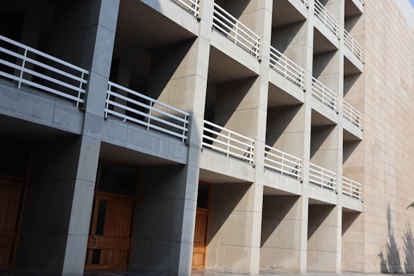 A modern architectural structure characterized by a grid of concrete modules, each with wooden doors and white railings, casting sharp shadows from sunlight.